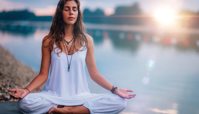 Beautiful young woman doing yoga exercise by the lake. Sitting in lotus position