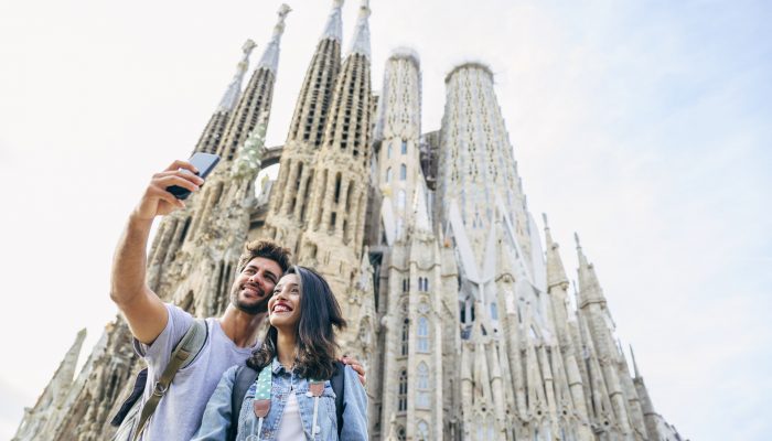 Low angle view of smiling male and female travelers enjoying city break in Barcelona and taking selfie with Sagrada Familia in background.